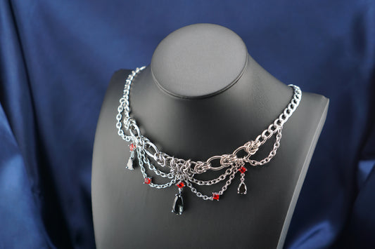 A silver-toned gothic-style chain necklace displayed on a matte black jewelry bust against a dark blue satin fabric background. The necklace features layered silver chains, a central cluster of silver links, small red bead accents, and dangling black teardrop and teardrop-shaped charms.