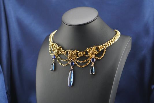 A gold-toned statement collar necklace with intricate chain links, draped chains, and three dangling teardrop blue gemstones, displayed on a black mannequin bust against a draped blue satin fabric background.