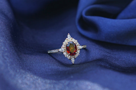 A delicate gold ring with a central red gemstone and small surrounding clear stones, resting on draped dark blue fabric.