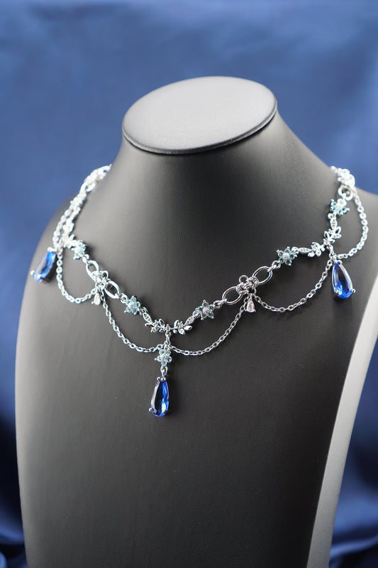 A silver-toned double-strand necklace with blue teardrop crystals and floral details, displayed on a matte black jewelry bust against a blue fabric background.
