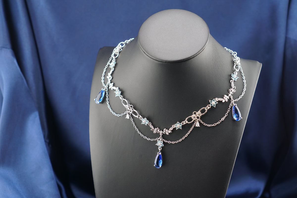 A silver-toned multi-strand necklace with blue teardrop crystals and small floral details, displayed on a black mannequin bust against a draped blue fabric background.