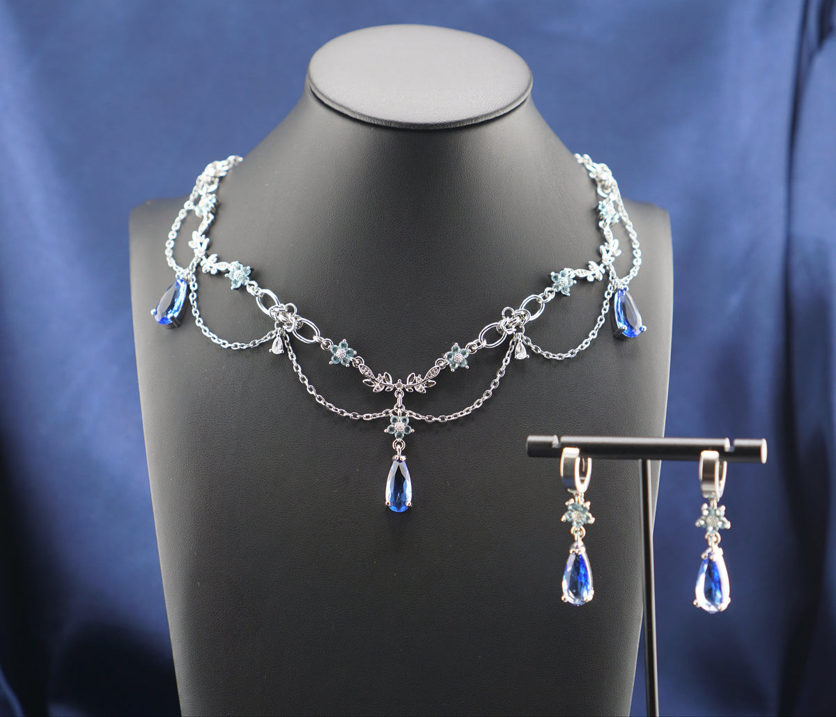 Silver-toned jewelry set featuring a layered chain necklace and matching drop earrings with blue teardrop crystals, displayed on a black mannequin bust against a blue satin fabric background.