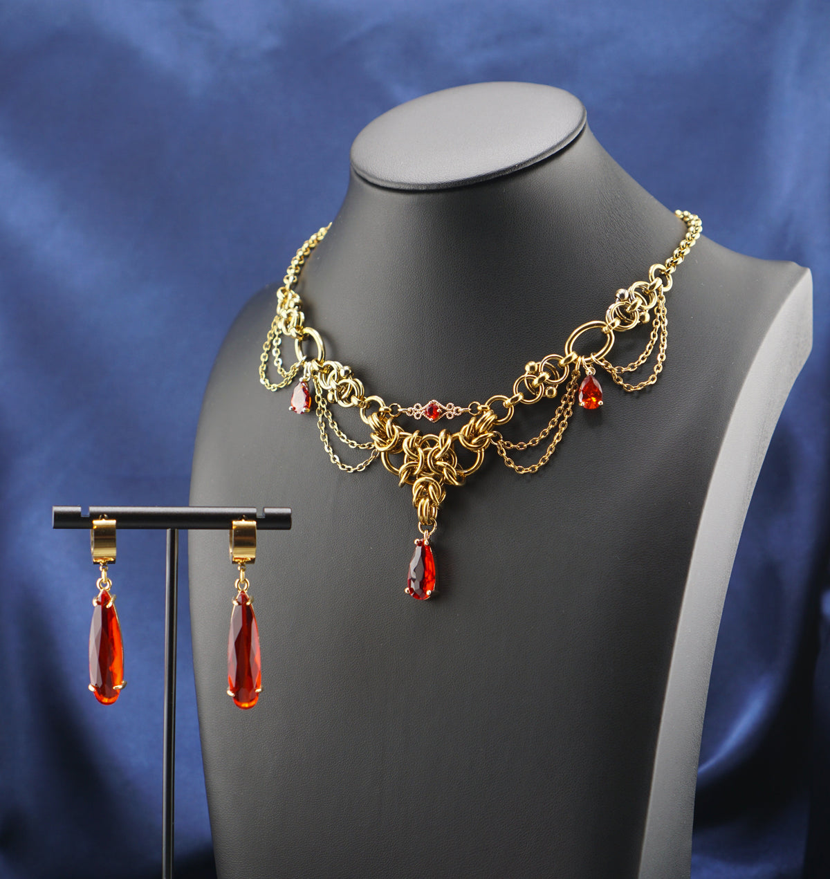 A gold-toned chain necklace and matching drop earrings displayed on a black mannequin bust, set against a draped blue satin fabric background.