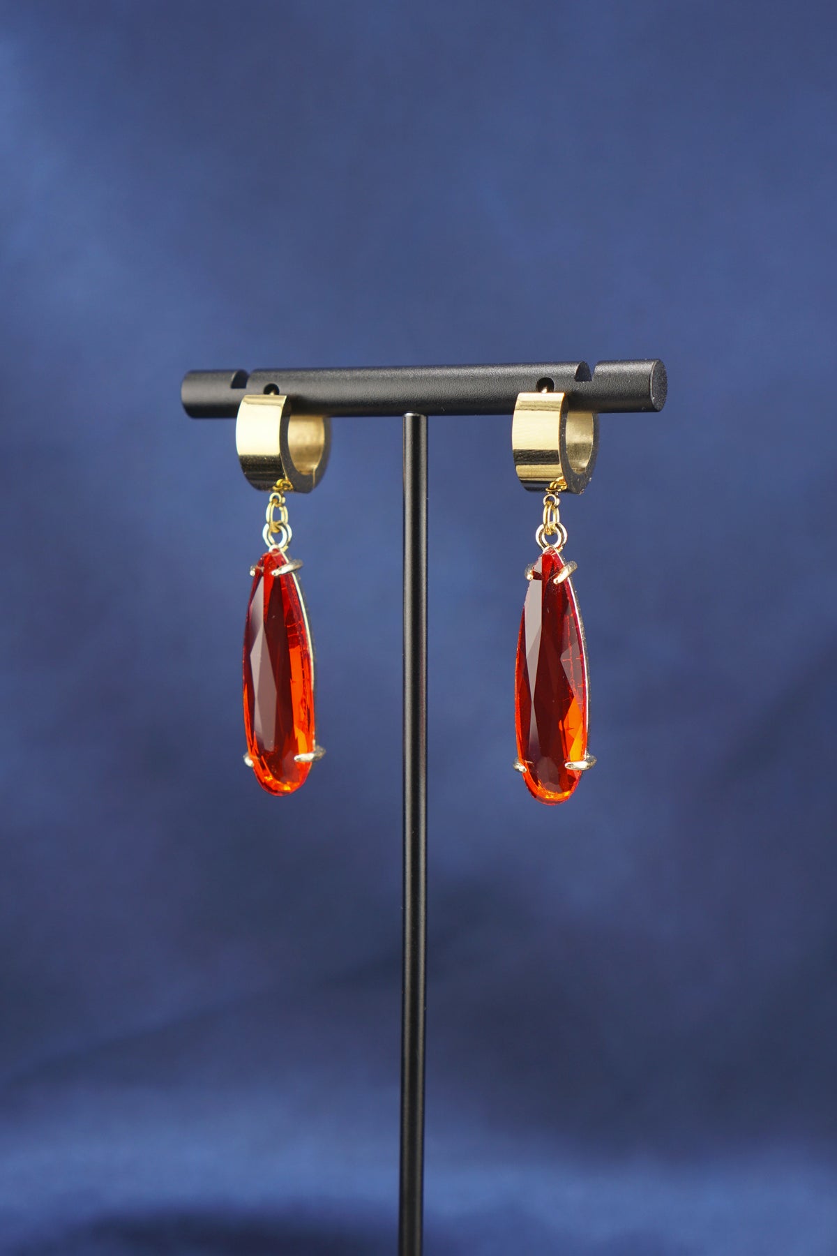 A pair of gold hoop earrings with faceted red teardrop drops, displayed on a black stand against a dark blue fabric background.
