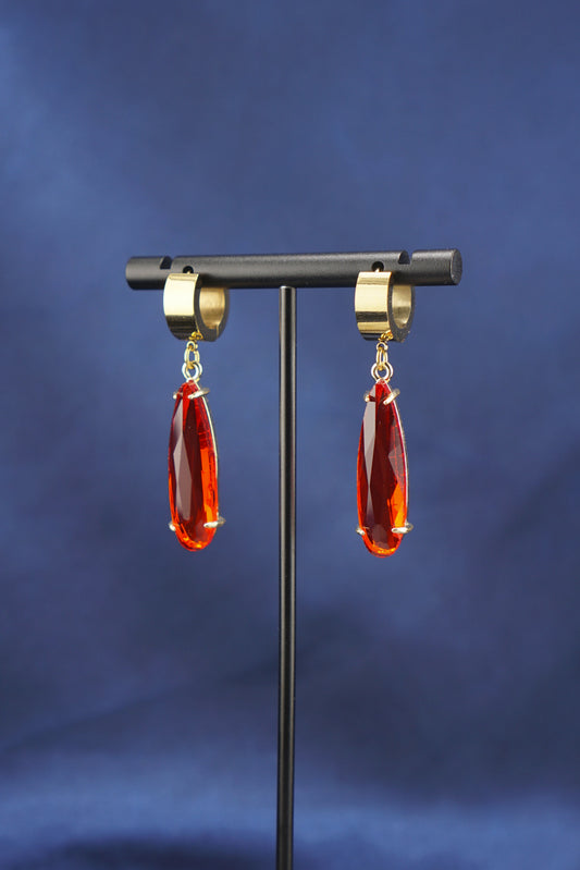 A pair of gold hoop earrings with faceted red teardrop drops, displayed on a black stand against a dark blue fabric background.