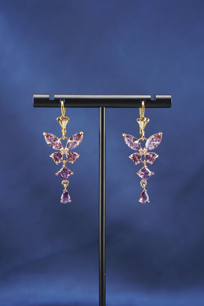 A pair of gold dangle earrings with purple gemstones arranged in a butterfly-like cluster, displayed on a black T-stand against a dark blue background.