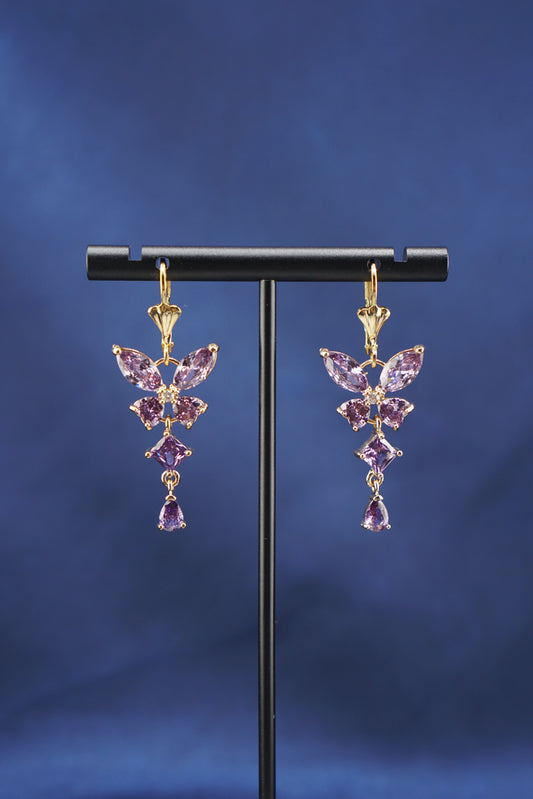 A pair of gold dangle earrings with purple gemstones arranged in a butterfly-like cluster, displayed on a black T-stand against a dark blue background.