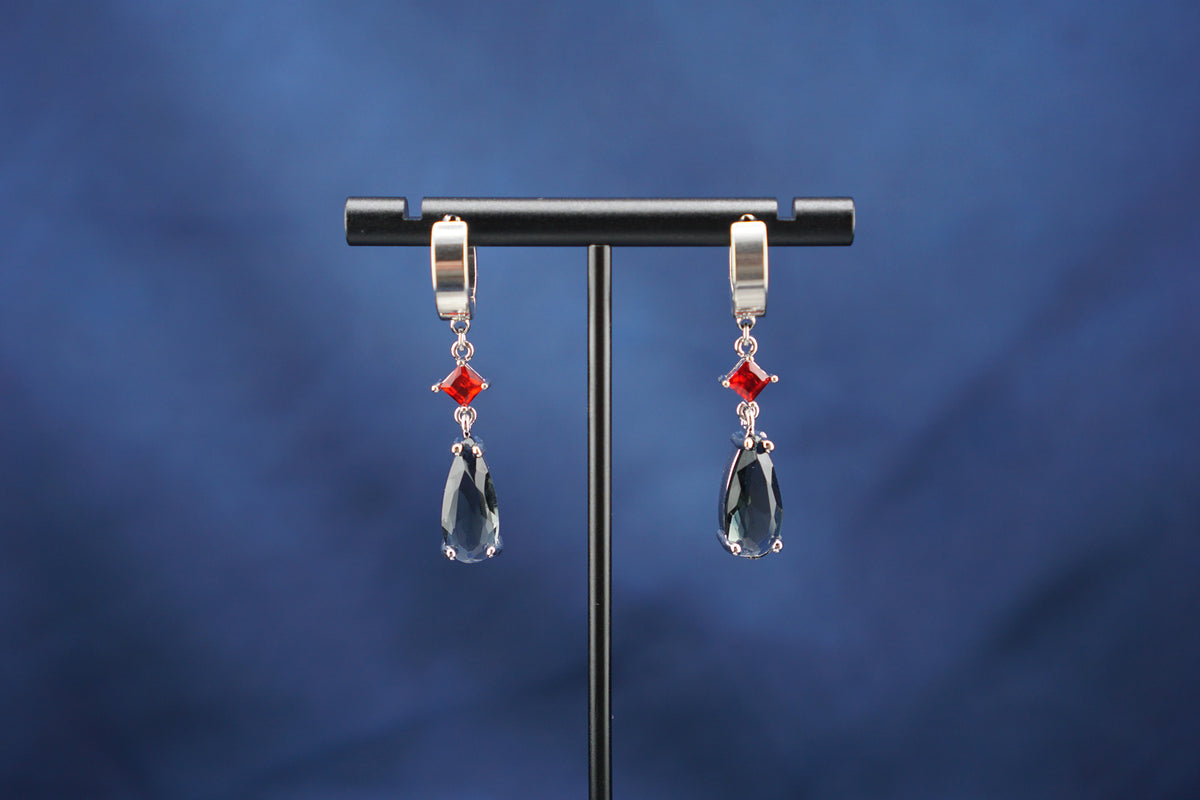 A pair of silver-toned drop earrings with small red gemstone accents and large clear teardrop crystals, hanging from a black display stand against a dark blue background.