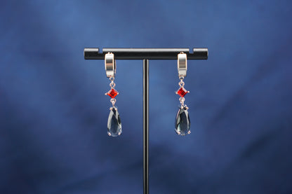 A pair of silver-toned drop earrings with small red gemstone accents and large clear teardrop crystals, hanging from a black display stand against a dark blue background.