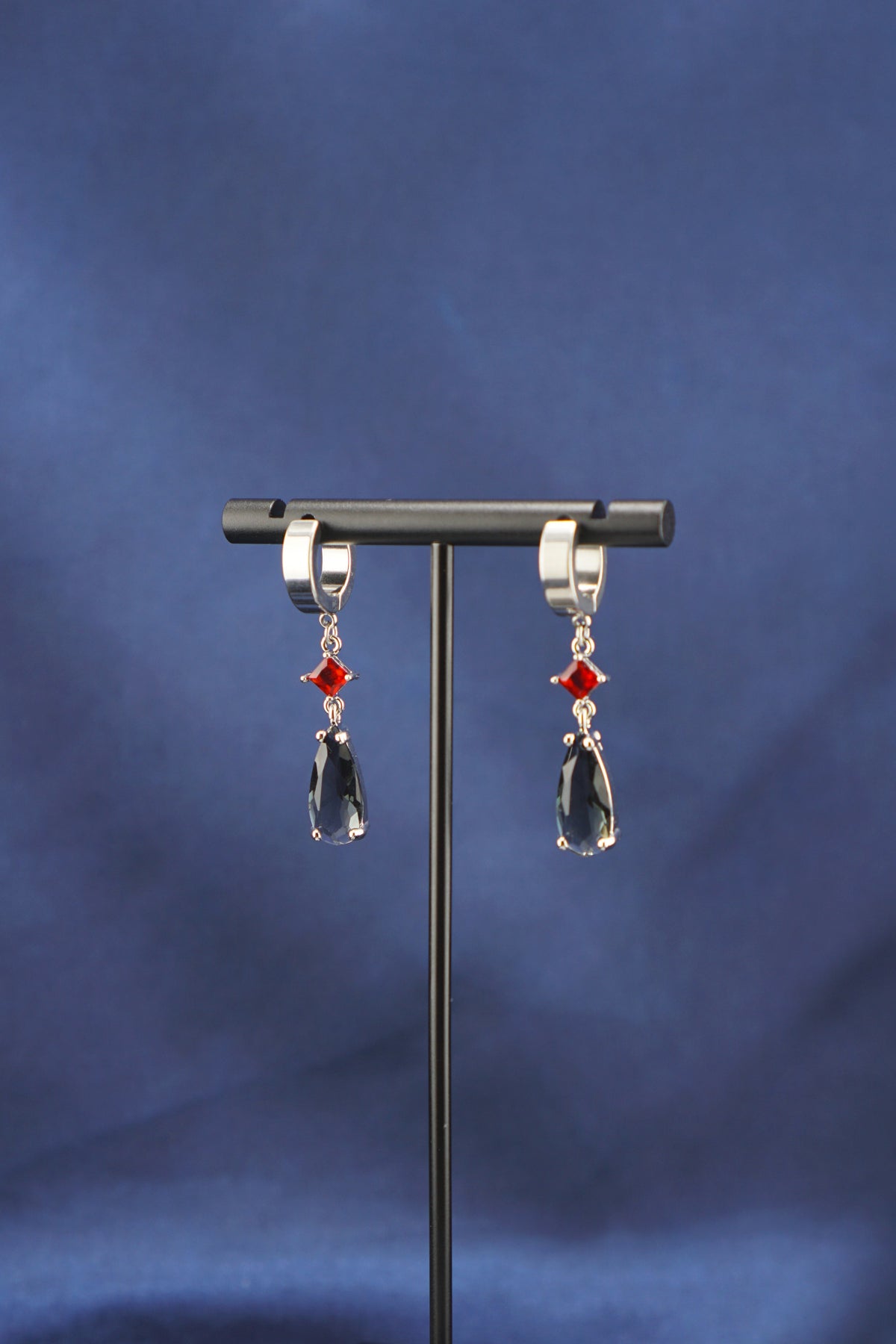 A pair of silver-toned drop earrings with red diamond-shaped accents and dark teardrop-shaped dangling charms, displayed on a black jewelry stand against a blue fabric backdrop.