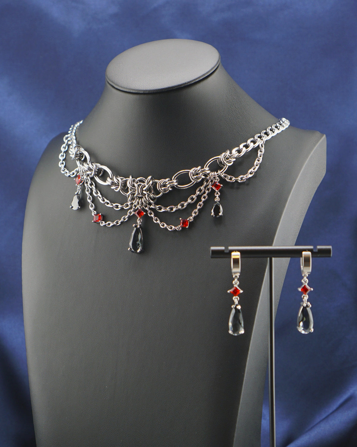 Silver-tone chain necklace and matching drop earrings with red bead accents, displayed on a black mannequin bust against a blue satin drape and white background.