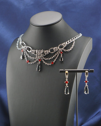 Silver-tone chain necklace and matching drop earrings with red bead accents, displayed on a black mannequin bust against a blue satin drape and white background.