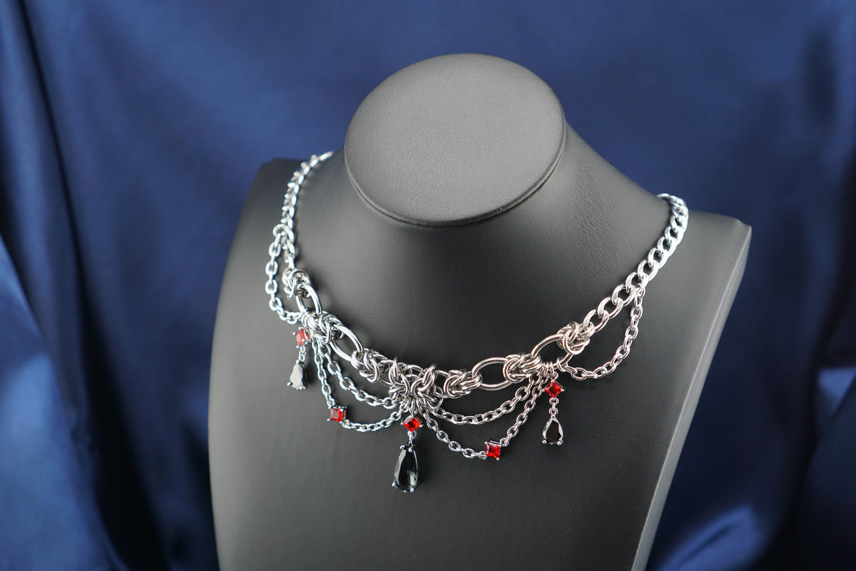 A silver-toned gothic-style chain necklace displayed on a matte black jewelry bust against a dark blue satin fabric background. The necklace features layered silver chains, a central cluster of silver links, small red bead accents, and dangling black teardrop and teardrop-shaped charms.