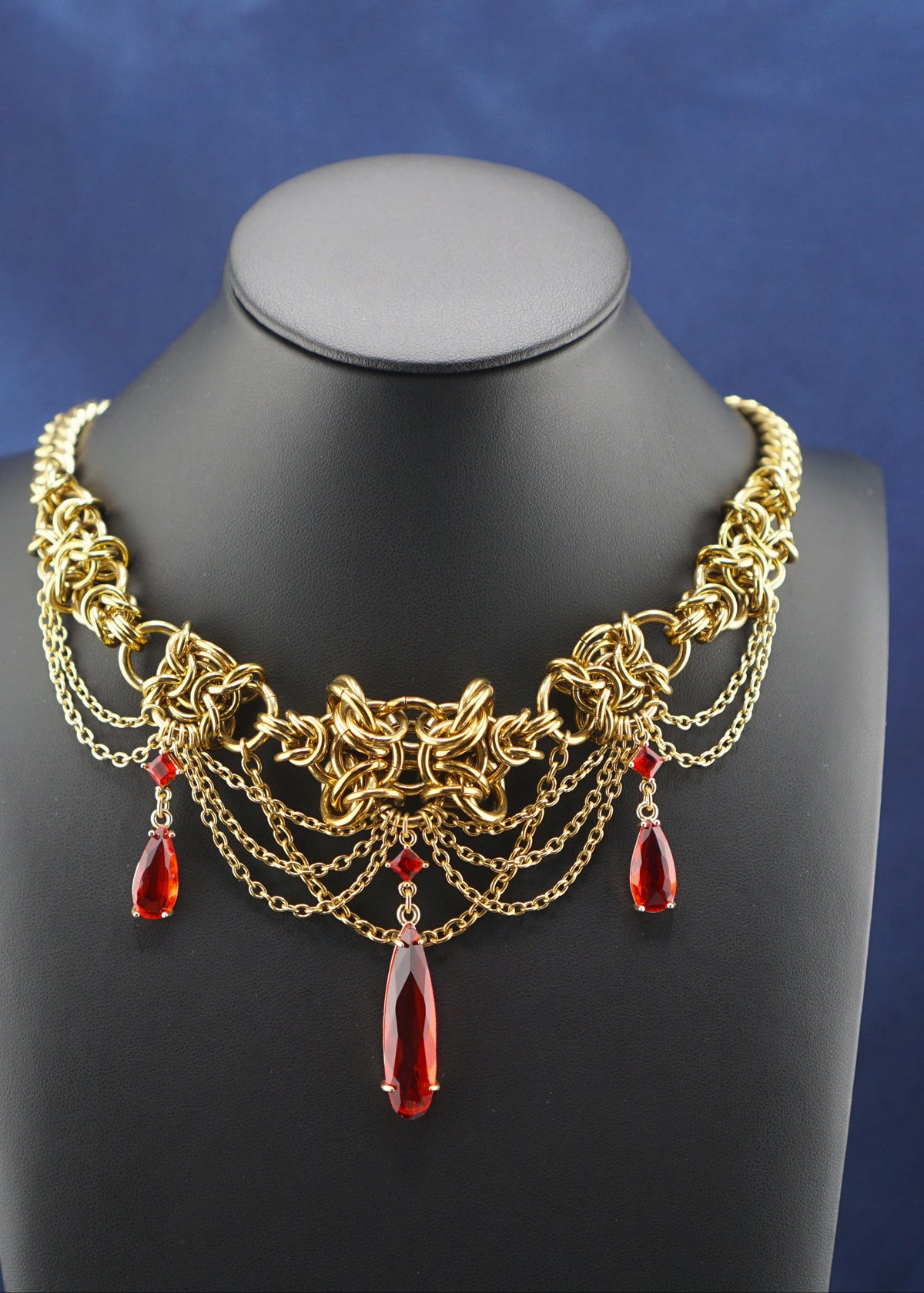 An ornate gold-toned statement necklace with red teardrop gemstones, displayed on a black mannequin bust against a draped blue satin fabric background.