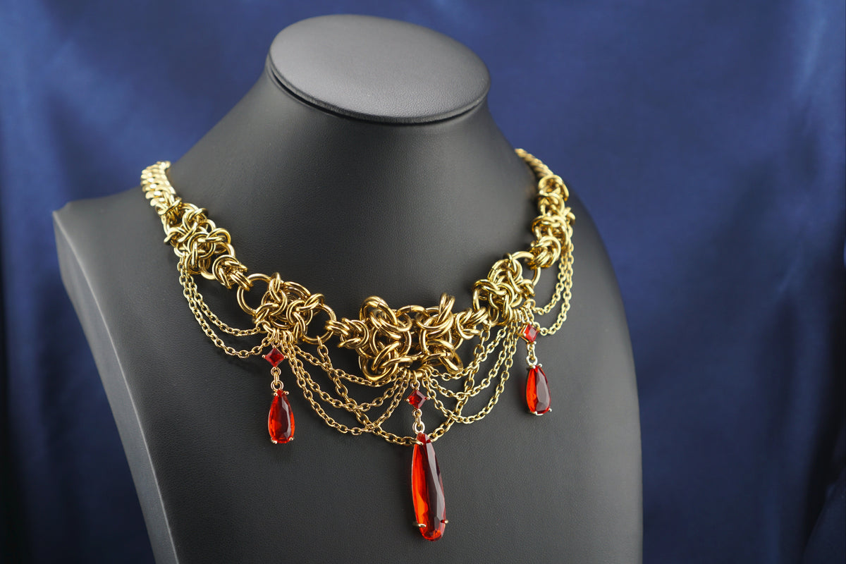 A gold-toned statement collar necklace with intricate chain and link details, accented by three dangling red teardrop beads, displayed on a black mannequin bust against a dark blue fabric backdrop.