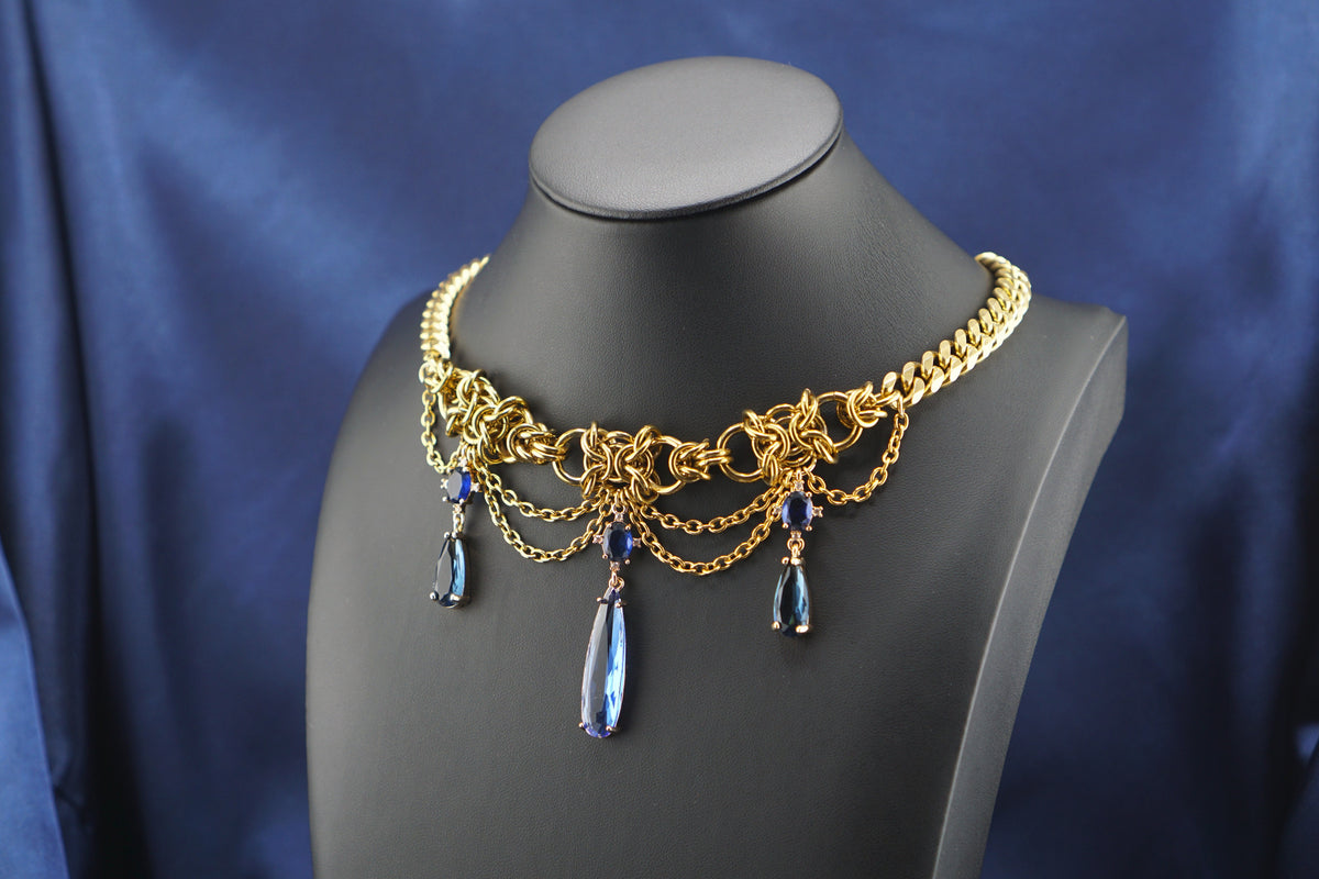 A gold-toned statement collar necklace with intricate chain links, draped chains, and three dangling teardrop blue gemstones, displayed on a black mannequin bust against a draped blue satin fabric background.