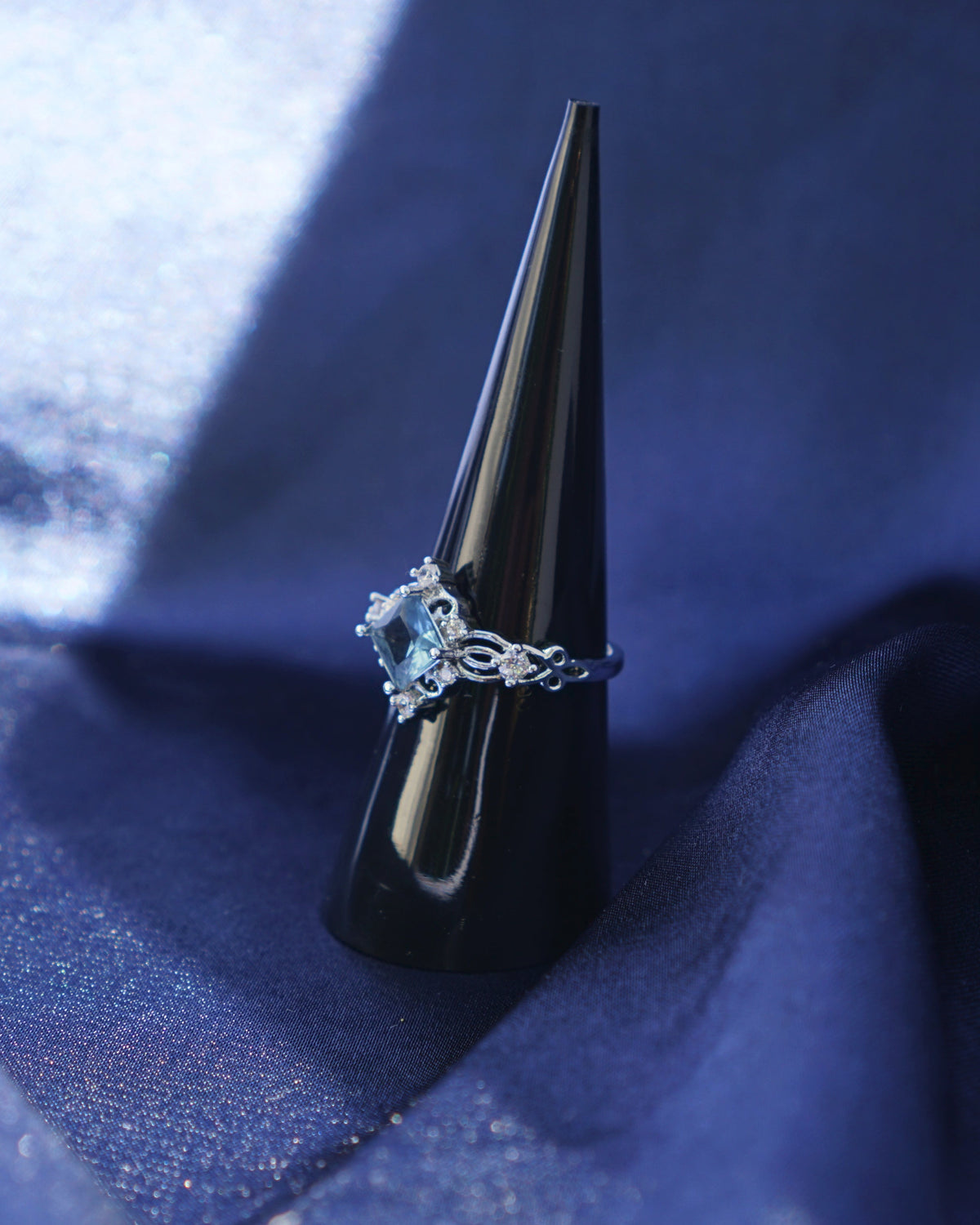 A silver ring with a light blue diamond-shaped gemstone displayed on a black cone ring holder, set against a draped dark blue fabric background.