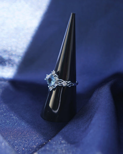 A silver ring with a light blue diamond-shaped gemstone displayed on a black cone ring holder, set against a draped dark blue fabric background.