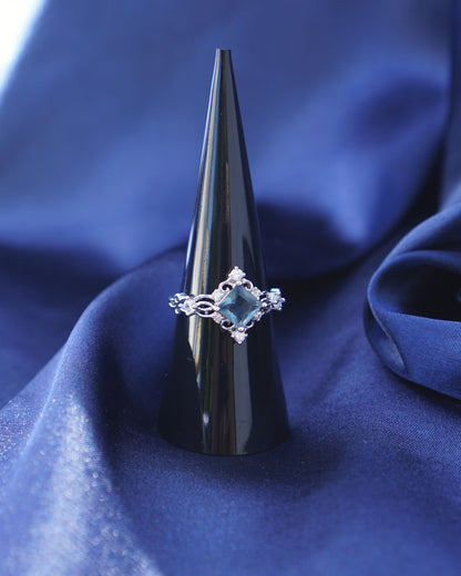 A silver ring with a central blue gemstone displayed on a black cone stand, set against a draped blue satin fabric background.