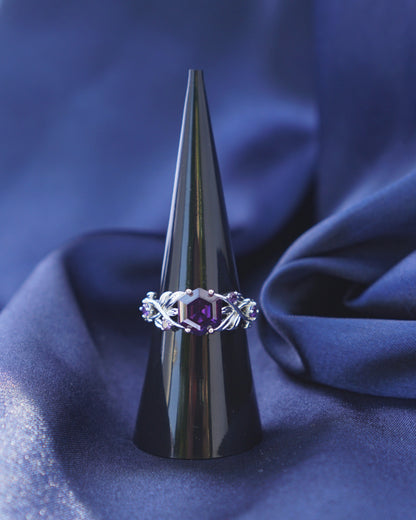 A silver ring with a central purple gemstone and decorative side stones, displayed on a black cone stand against a draped blue fabric background.