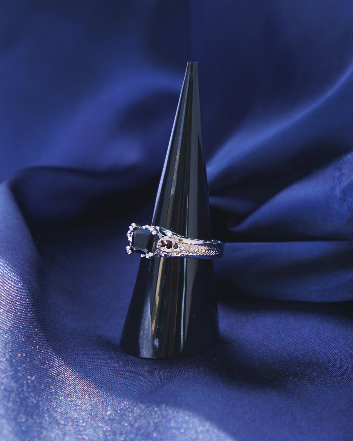 A silver ring with a black onyx gemstone displayed on a black cone stand, nestled in dark blue draped fabric.