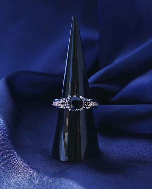 A silver ring with a square-cut clear gemstone displayed on a black cone stand, set against draped dark blue fabric.