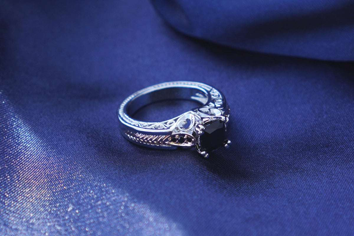 A silver-toned ring with a onyx square gemstone rests on draped navy blue fabric