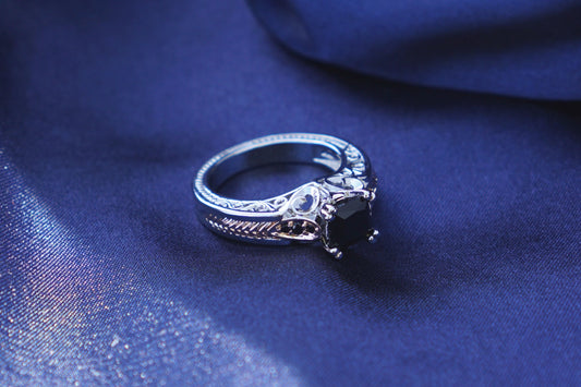 A silver-toned ring with a onyx square gemstone rests on draped navy blue fabric