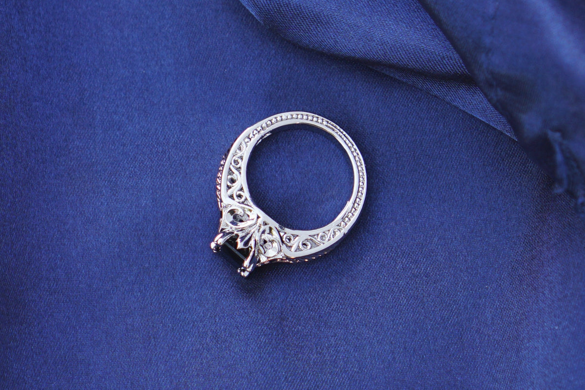 A silver ring with a black onyx gemstone and a fleur de lys design, set with small onyx stones and a rose gold accent, resting on draped royal blue satin fabric.