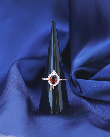 A close-up of a gold ring with a star-shaped setting and a central red gemstone surrounded by small clear stones, displayed on draped royal blue fabric.