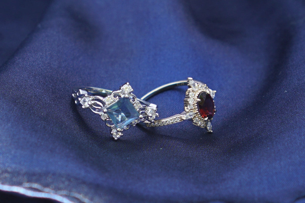 A silver-toned ring with blue gemstone and garnet and gold toned ring rests on draped dark blue fabric