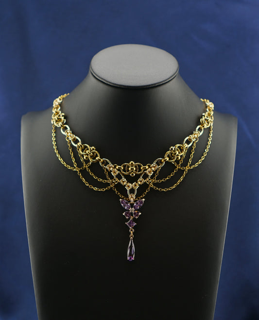 A gold-toned multi-strand chain necklace with a purple crystal pendant, displayed on a black mannequin bust against a blue satin fabric backdrop.