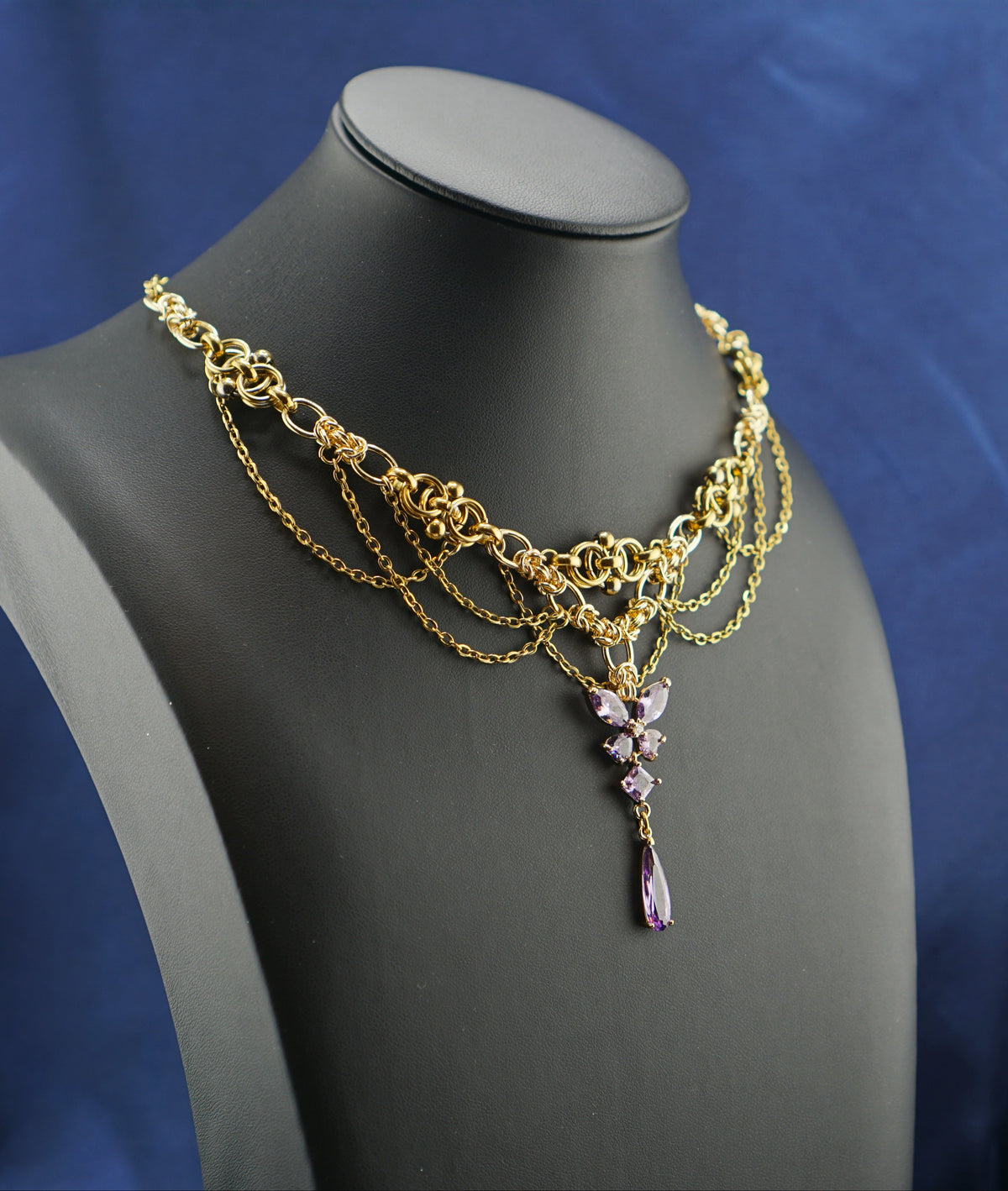 A gold-toned multi-strand chain necklace with a purple crystal pendant, displayed on a black jewelry bust against a blue satin fabric background.
