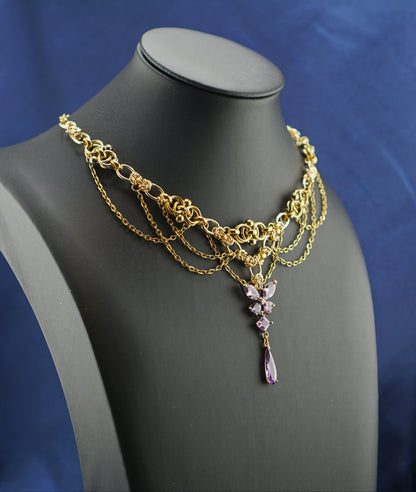 A gold-toned multi-strand chain necklace with a purple crystal pendant, displayed on a black jewelry bust against a blue satin fabric background.