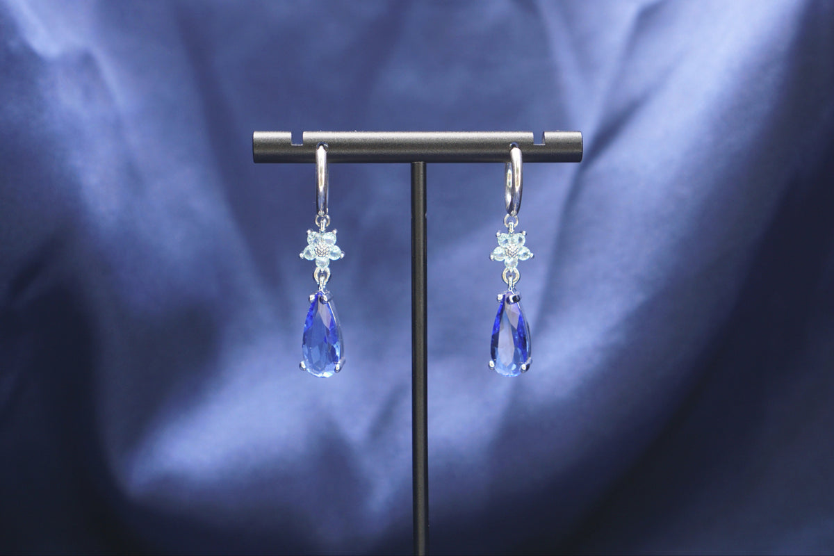 A pair of silver-toned dangle earrings with clear crystal starburst charms and blue teardrop drops, displayed on a black T-stand against a draped blue fabric background.