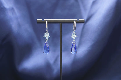 A pair of silver-toned dangle earrings with clear crystal starburst charms and blue teardrop drops, displayed on a black T-stand against a draped blue fabric background.