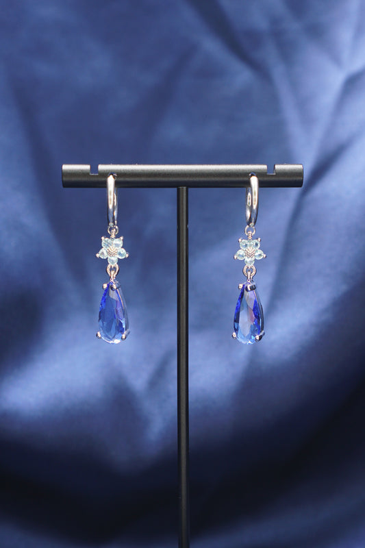 A pair of silver-toned dangle earrings with clear faceted stones in a floral setting and blue teardrop-shaped drops, displayed on a black stand against a draped blue fabric background.